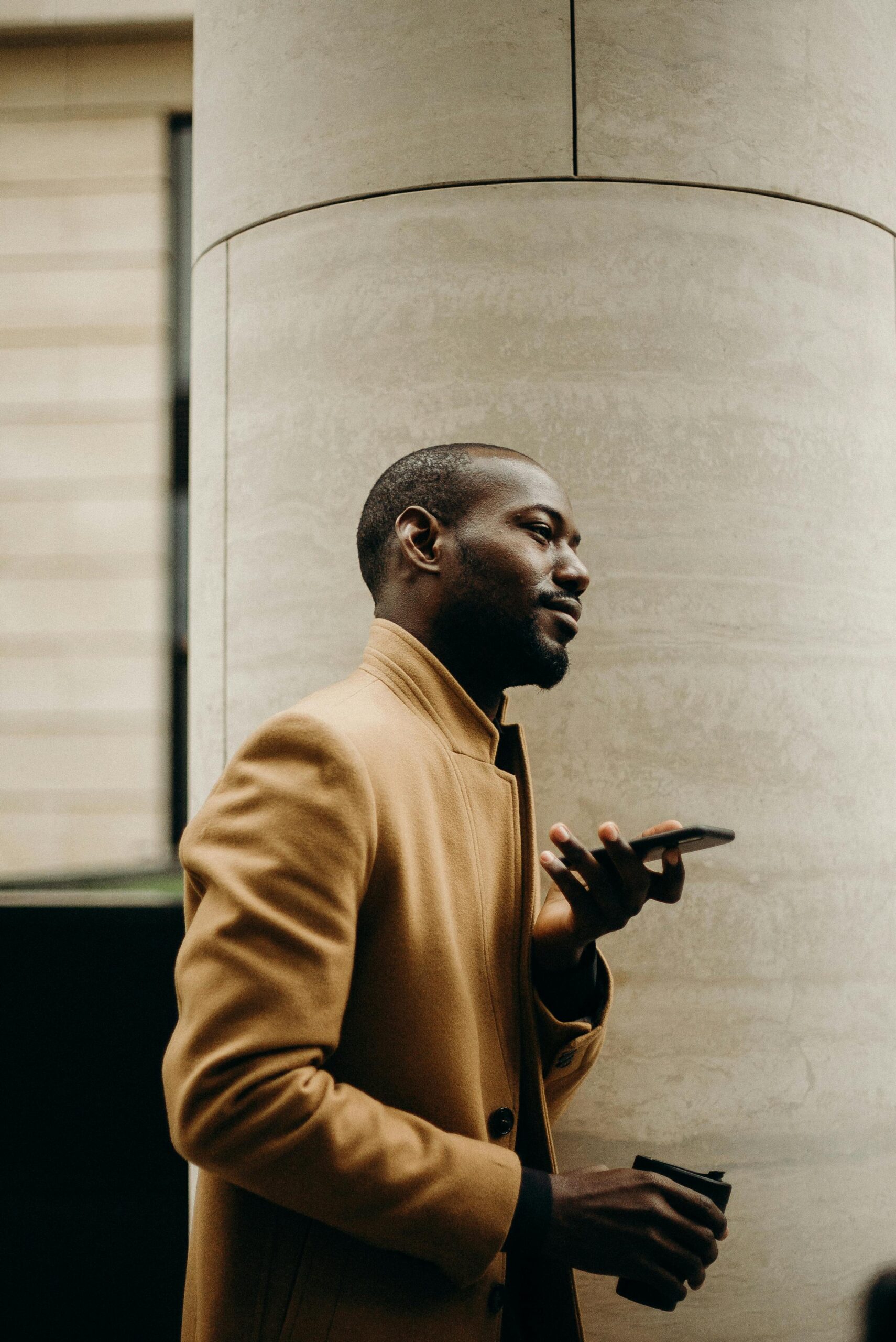 Side view of a stylish black man in a trench coat talking on his smartphone outdoors.